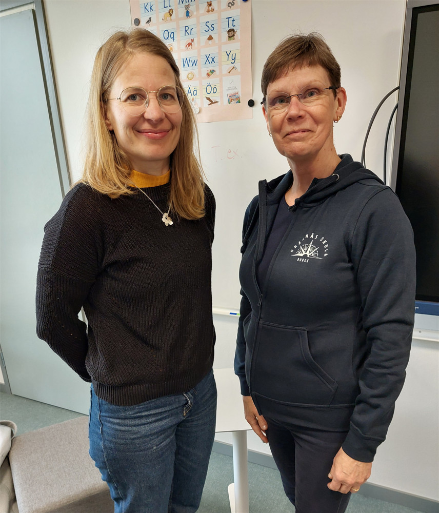 På bilden syns två medelålders kvinnor som står i ett klassrum. Man ser en ljusblå dörr, en vit vägg och en plansch med bilder samt en del av en svart skärmtill höger.Kvinnan till vänster har axellångt ljust hår, diskreta glasögon, en svart tröja ovanpå en gul blus och jeans. Hon ser rakt in i kameran och ler med stängd mun. Kvinnan till höger har kortklippt brunt hår, diskreta glasögon och en huvtröja och byxor i mörkblått. Hon ser också rakt in i kameran och ler.