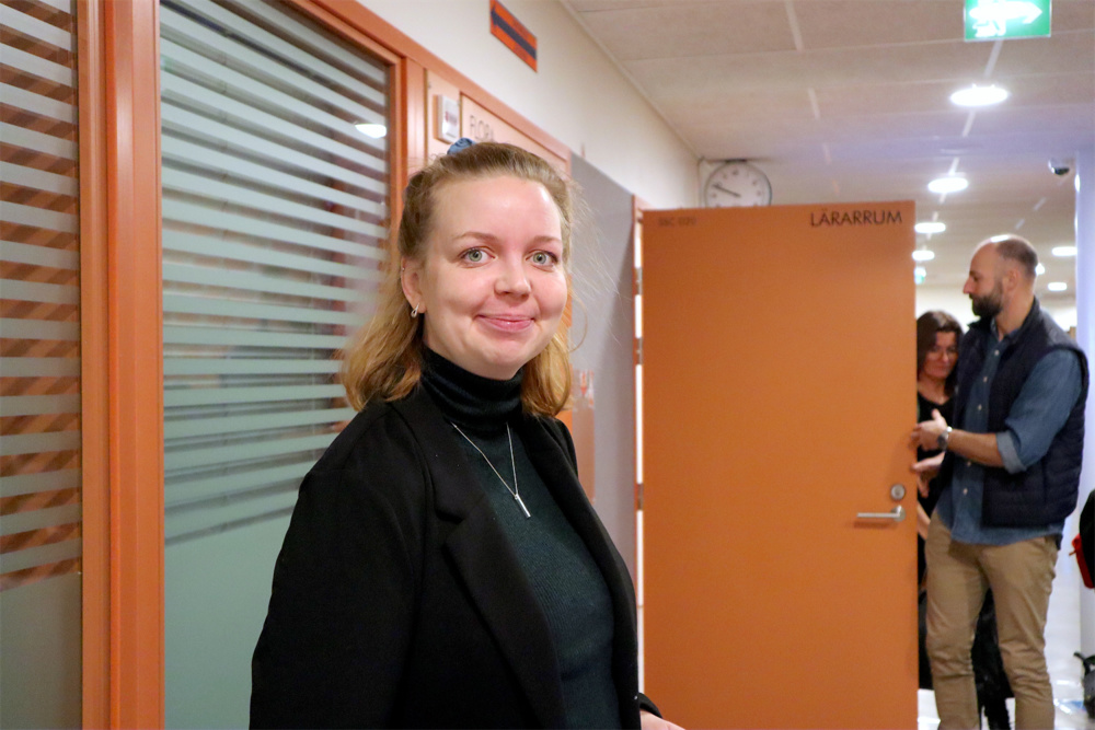 På bilden syns en medelålders kvinna soms tår i en skolkorridor med ljusa väggar och orangea dörrar och fönster med orangea karma. I bakgrunden syns också en klocka och elever . Kvinnan har medellångt ljust hår som är uppsatt på huvudet. Hon har en mörkblå tröja och tittar in i kameran och ler.